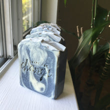 Load image into Gallery viewer, Natural green and black swirled soap sitting on a white window sill in the natural light. The soap is stamped with "Soaps for Change" and a houseplant is sitting behind it.