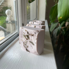 Load image into Gallery viewer, Handmade soap bars sitting upright on a well-lit window sill. The soap is light-purple and square-shaped, topped with herbs and dried botanicals.