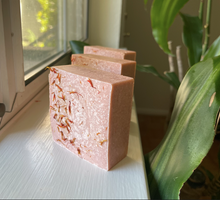 Load image into Gallery viewer, Three peach colored, square-shaped soap bars on a window sill topped with red safflower petals, with a green plant in the background