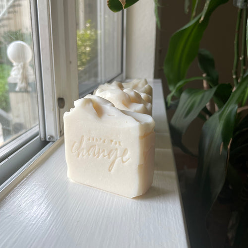 Handmade soap bars sitting upright on a well-lit window sill. Soaps are naturally off-white colored. There are plants in the background.