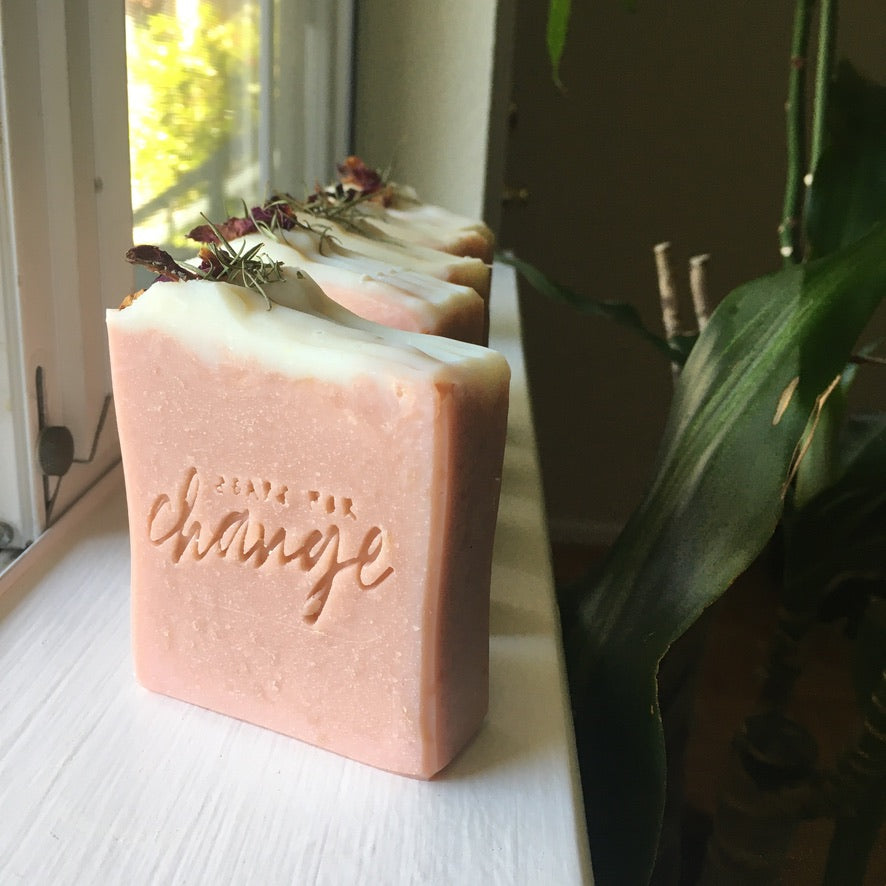 A pink soap topped with a small white layer of soap as well as rose petals and evergreen needles, sitting on a white window sill in the natural light. The soap is stamped with "Soaps for Change" and a houseplant is sitting behind it.