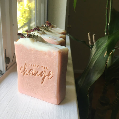 A pink soap topped with a small white layer of soap as well as rose petals and evergreen needles, sitting on a white window sill in the natural light. The soap is stamped with 