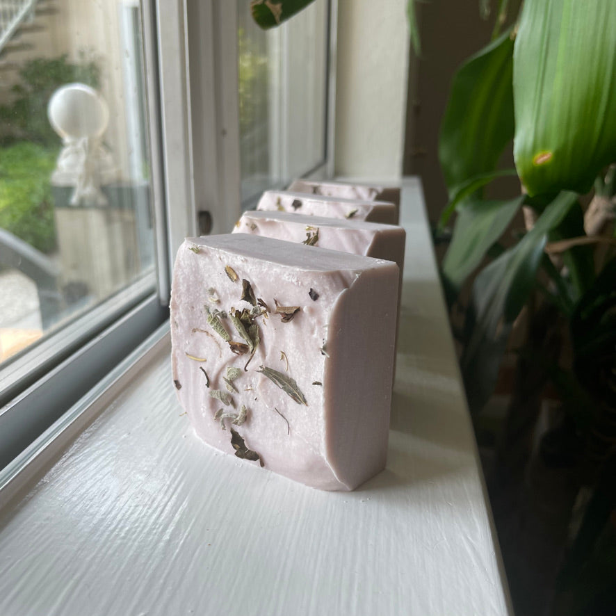 Handmade soap bars sitting upright on a well-lit window sill. The soap is light-purple and square-shaped, topped with herbs and dried botanicals.