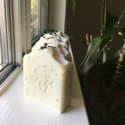 A natural colored off-white soap with black specks sitting on a white window sill in the natural light. The soap is stamped with 