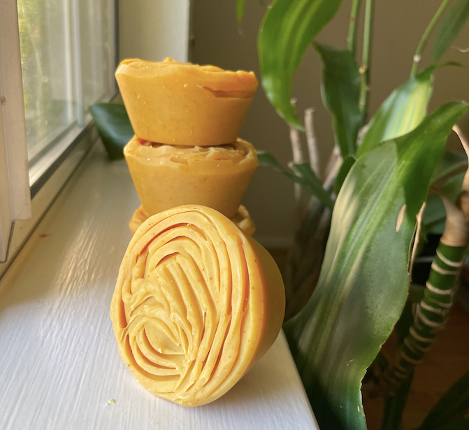 Circular orange soaps stacked on a window sill. The top of the soap has a design carved with a fork. There is a green houseplant in the background.
