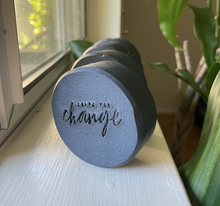 Load image into Gallery viewer, Four black, circular soap bars sitting on a window sill in natural light. The soaps are stamped with &quot;Soaps for Change&quot; and there is a green houseplant in the background.
